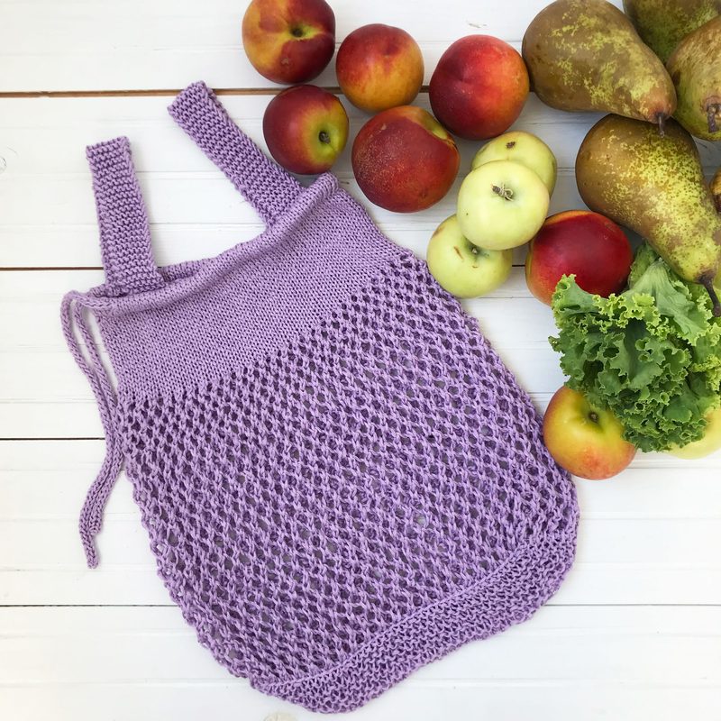 A knitted produce bag in light lavender rests on a white wooden surface, featuring a netted design with two sturdy straps. Surrounding the bag are various fruits, including red apples, green apples, and brown pears, along with a bunch of leafy greens, creating a vibrant, natural display. The overall composition showcases the produce bag and its intended use for carrying fresh items.