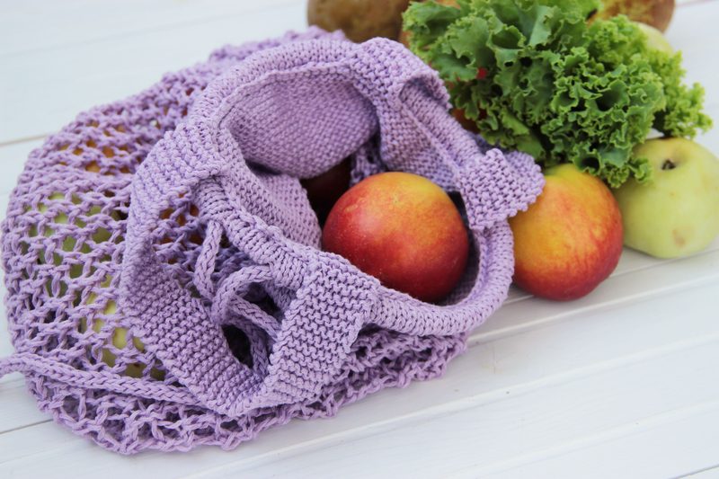 A lavender knitted mesh produce bag sits on a light wooden surface, partially filled with three red apples and surrounded by fresh green lettuce leaves and a potato. The bag's open design showcases its knitted texture and allows visibility of the contents inside.