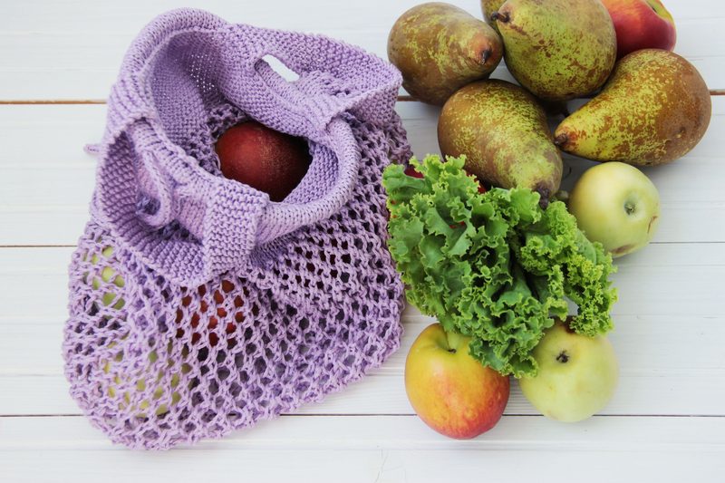 A handmade purple knitted produce bag sits on a white wooden surface, partially filled with red and green apples. Surrounding the bag are several green and brown pears, as well as leafy green lettuce and additional apples, creating an arrangement of fresh produce. The bag features an open weave design, showcasing the items inside. The overall composition is bright and colorful, highlighting the contrast between the knitted texture and the smooth surface of the fruits and vegetables.