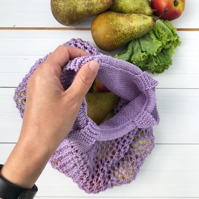 A hand holds a light purple knitted produce bag, which has a lace pattern and is partially open. Inside the bag, several fruits and vegetables are visible, including two green pears, a red apple, and some leafy lettuce. The background features a light wooden surface, creating a clean, bright look.