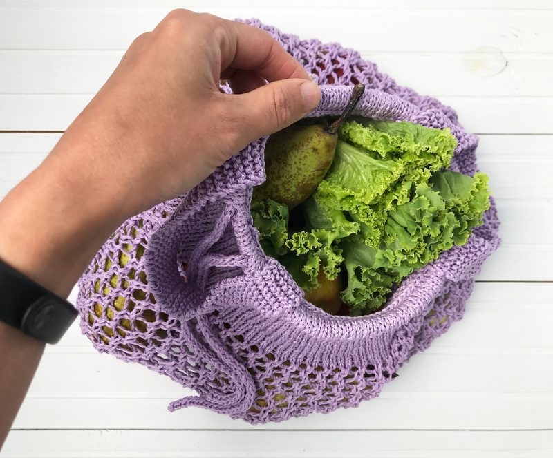 A hand holds the top edge of a lilac-colored knitted produce bag filled with green lettuce and pears. The bag features an open, airy knit pattern and is positioned against a white wooden surface, emphasizing the texture and color of both the bag and its contents.