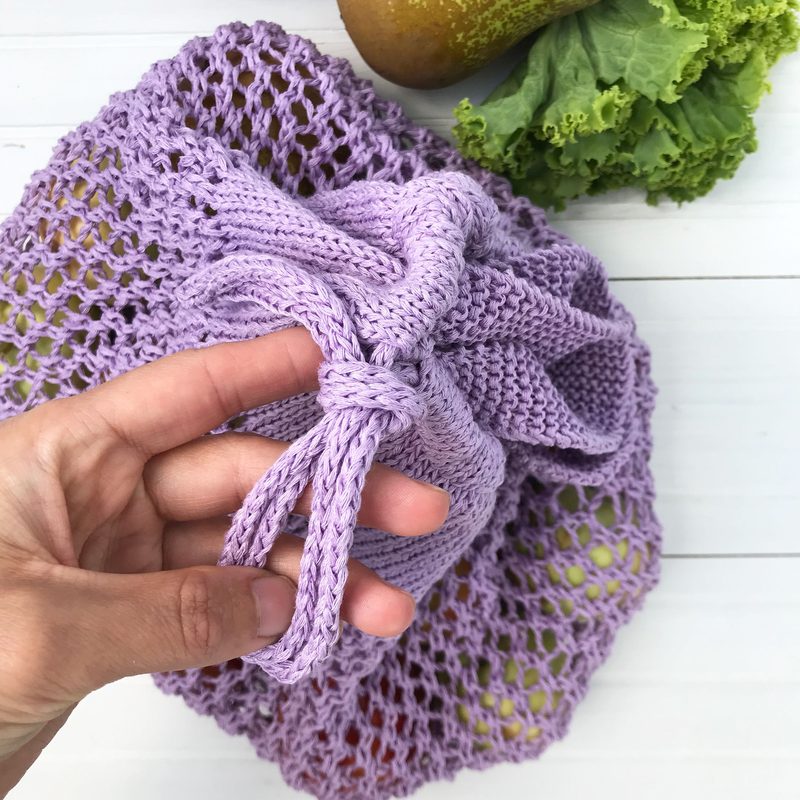 A close-up image of a lavender knitted produce bag, partially closed with a knot at the top. The bag is made of a loose, net-like pattern, allowing some visibility of items inside. A person's hand is holding the bag, and a green leafy lettuce and a pear are partially visible next to it, resting on a white wooden surface.
