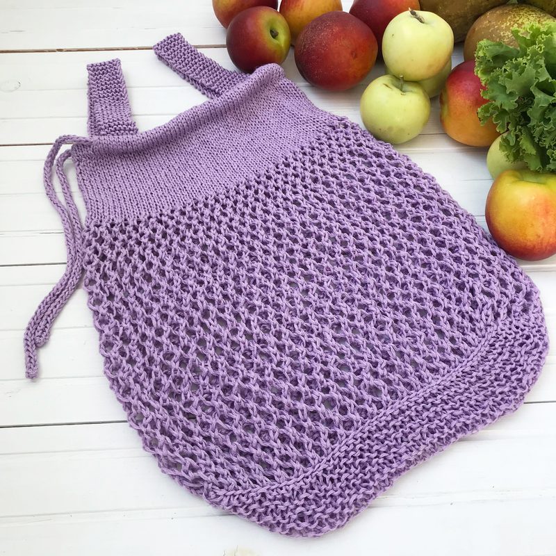 A lavender knitted produce bag is displayed on a white wooden surface, featuring a netted lower section and two straps at the top. Surrounding the bag are various fruits and vegetables, including red and green apples and leafy lettuce, creating a colorful contrast with the bag. The textures of the knitted fabric and the produce are clearly visible against the simple background.