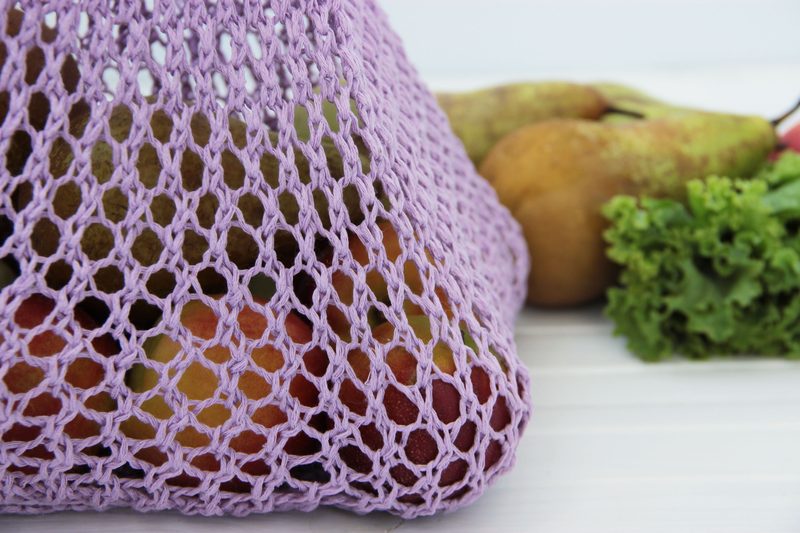 A knitted produce bag in light purple filled with various fruits, including apples and pears, alongside a piece of lettuce. The bag's mesh pattern is clearly visible, showcasing the contents. The background is simple and light-colored, enhancing the prominence of the bag and its contents.