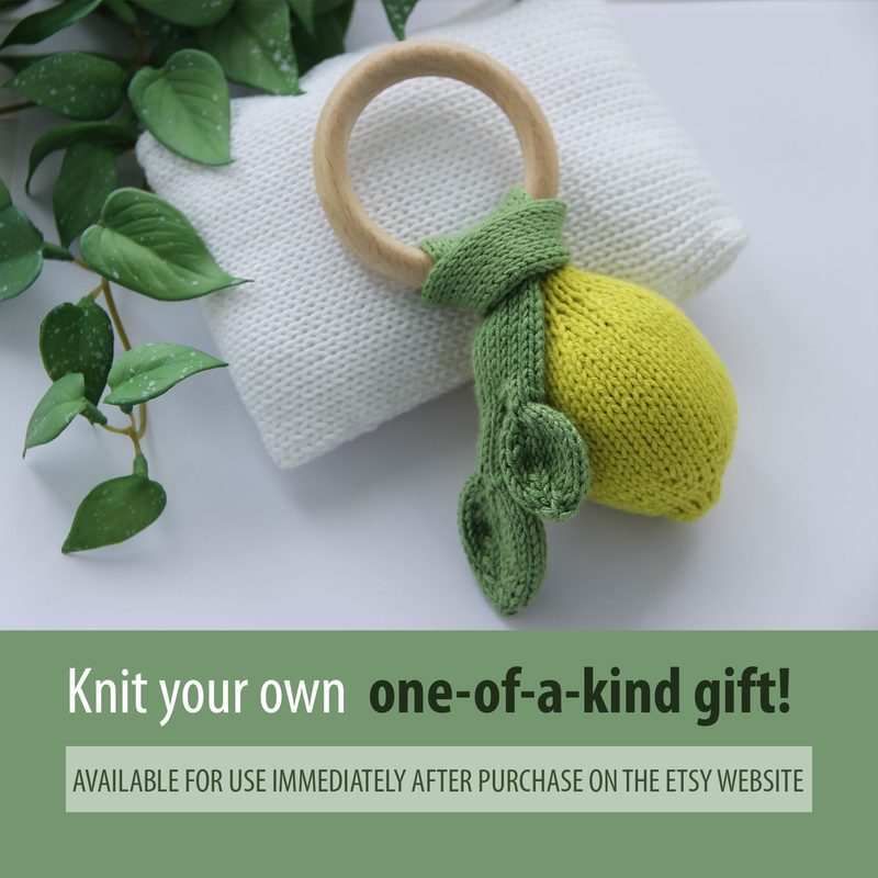 A hand-knitted lemon toy is displayed on a white textured fabric, resting beside a wooden teething ring. The lemon is bright yellow with a knitted green leaf and a knitted green tail. The background features a few green leaves, adding a natural element to the scene. The composition is centered and showcases the vibrant colors of the lemon and the contrasting textures of the fabric and wood.
