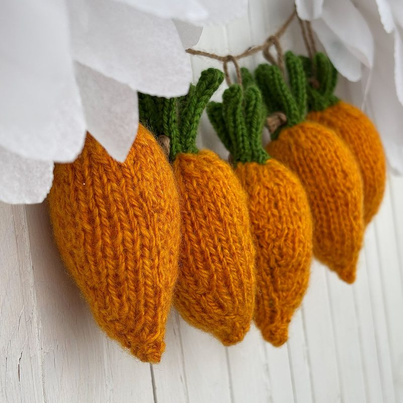 The image displays several knitted mini carrots in a row, showcasing a bright orange color with a textured knit pattern. Each carrot features green knitted leaves at the top. The background is light and neutral, enhancing the vibrant carrots, and there are soft white flowers subtly framed at the top, adding contrast to the bright colors of the carrots. The overall composition is visually appealing and arranged in a decorative manner.