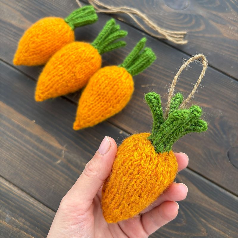A hand holds a knitted mini carrot, which is bright orange with green knitted leaves at the top. In the background, three identical mini carrots are arranged on a wooden surface. The overall color palette features vibrant orange and green against the warm brown tones of the wood. The carrots are shown in a close-up view, highlighting the texture of the knitting. A piece of twine is visible, suggesting they may be used for decoration.