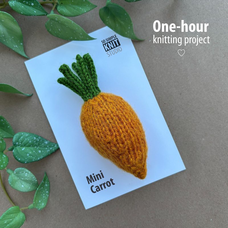 The image features a small, knitted mini carrot positioned on a white card. The carrot has a vibrant orange body with a textured finish, and it is topped with green yarn leaves that are neatly styled. The card includes the text "Mini Carrot" at the bottom and "One-hour knitting project" in the upper right corner. Surrounding the card are green plant leaves, providing a natural background and a pop of color.