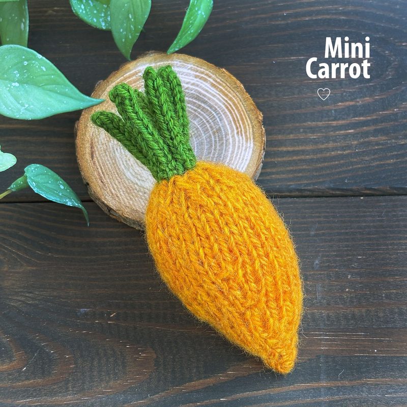 A knitted mini carrot, featuring a bright orange body with a textured surface and green leaf accents at the top. The carrot is placed on a circular wooden slice, which is set against a dark wooden background. There's a label to the right that reads "Mini Carrot" in a simple font. Green leaves are partially visible on the left side of the image, adding a natural touch to the scene.