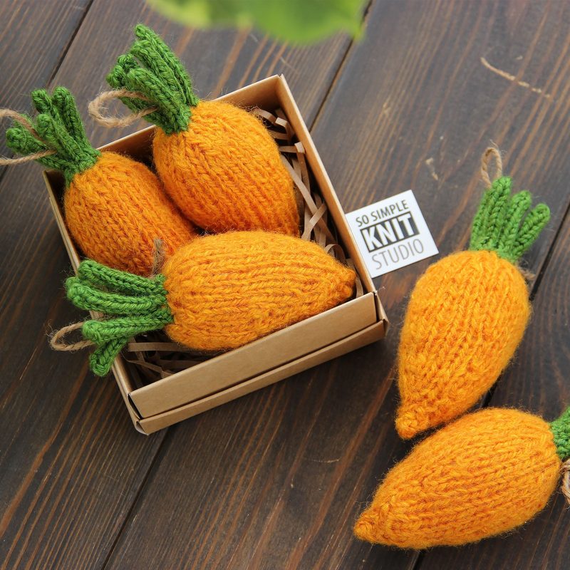 A small, square cardboard box filled with four knitted mini carrots, featuring bright orange bodies and green leafy tops. The carrots are arranged in a natural, casual style, with visible texture from the yarn. The box is placed on a rustic wooden surface, and a small label in the box reads "SO SIMPLE KNIT STUDIO." The background is softly blurred, focusing attention on the box and the carrots.