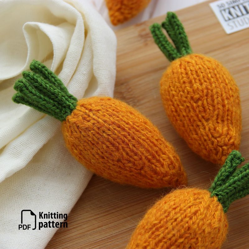Three mini knitted carrots with a vibrant orange body and green leafy tops are arranged on a wooden surface. The carrots are textured, showcasing a knitted pattern. A piece of cream-colored fabric is draped nearby, adding contrast to the warm colors of the carrots. In the bottom left corner, there is a graphic symbol indicating it is a PDF knitting pattern. The overall composition is brightly lit, highlighting the details in the knitting.