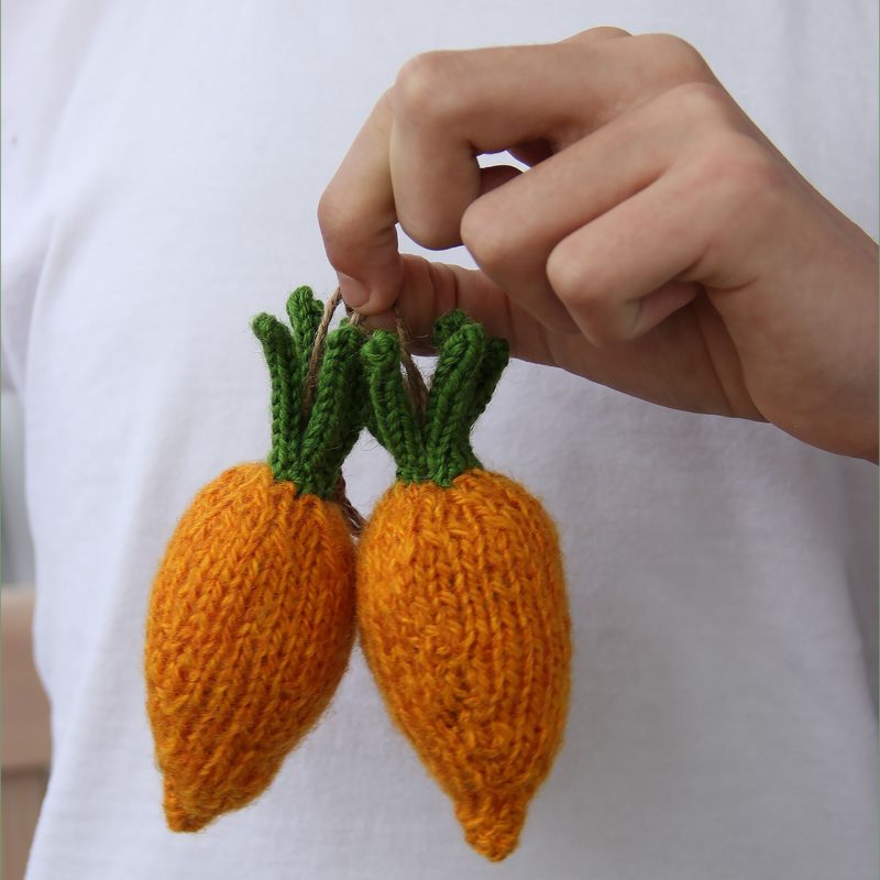 A person's hand holds two knitted mini carrots, both in vibrant orange with green leafy tops. The carrots are textured with visible stitches, and the hand is partially in view, with a plain white background suggesting a casual setting. The focus is on the knitted details of the carrots.