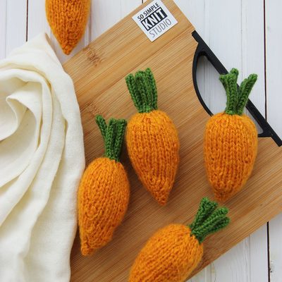 The image features five knitted mini carrots arranged on a wooden cutting board, with three carrots placed upright and two lying on their sides. The carrots are bright orange with green knitted tops. A cream-colored, textured fabric is draped casually beside the cutting board. The background is a light, wooden surface, adding contrast to the vibrant colors of the carrots. The composition is well-lit, emphasizing the intricate knitting detail.