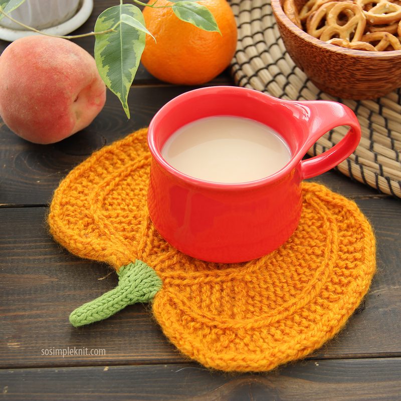 The image shows a vibrant orange pumpkin-shaped knitted coaster with a green knitted stem, placed on a wooden surface. A bright red mug filled with a light-colored drink sits on the coaster. Surrounding the mug are a peach, an orange, and a bowl of pretzels, creating a cozy, seasonal setting. A few green leaves are partially visible in the background, adding to the natural feel of the composition.
