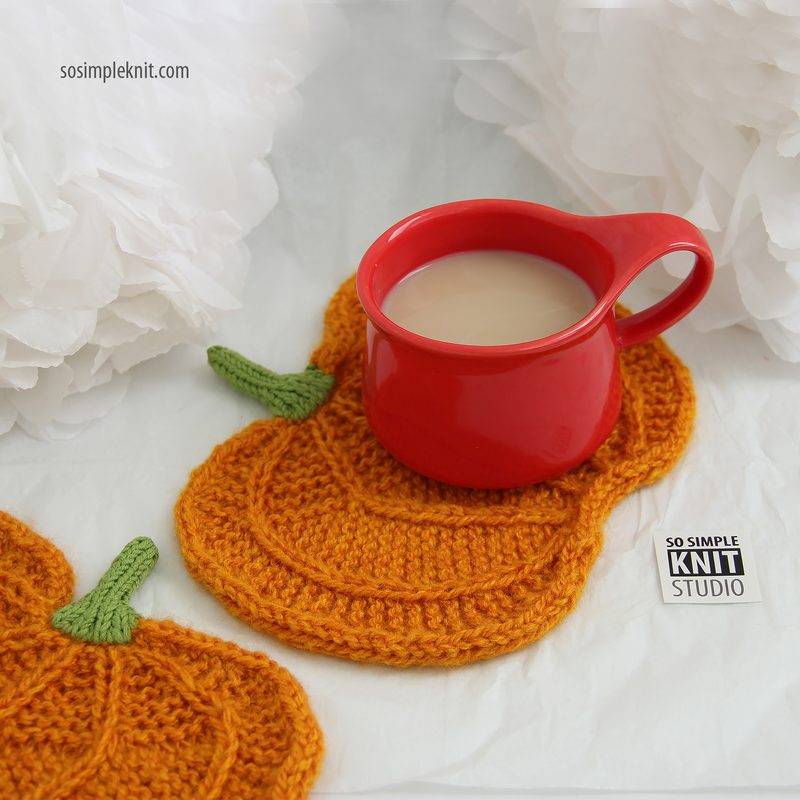 The image features a knitted pumpkin coaster in vibrant orange, shaped like a pumpkin with a green stem, resting on a soft white background. A small red ceramic cup filled with a light-colored beverage sits on top of the coaster, creating a cozy scene. Fluffy white tissue paper decoratively frames the composition, enhancing the softness of the overall aesthetic. A logo for "So Simple Knit Studio" is visible near the bottom right corner.