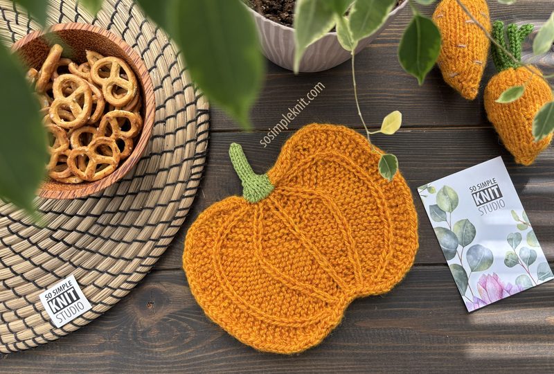 A knitted pumpkin coaster in vibrant orange is placed on a dark wooden table alongside a woven placemat and a small bowl of pretzels. Nearby, a printed knitting pattern brochure featuring floral illustrations is visible, and a small tag with "So Simple Knit Studio" is displayed. Green leaves from a plant hang slightly over the scene, adding a natural touch.