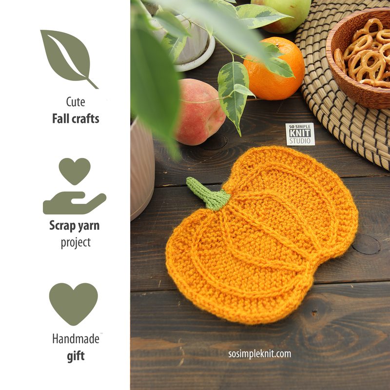 The image features a knitted pumpkin coaster in vibrant orange, positioned on a dark wooden table. The coaster is detailed with textured stitching and a small green stem at the top. Surrounding the coaster are natural elements, including a woven basket filled with pretzels and fresh fruits like apples, with a few green leaves visible. The overall composition has a warm, autumnal feel, emphasizing a cozy crafting theme.