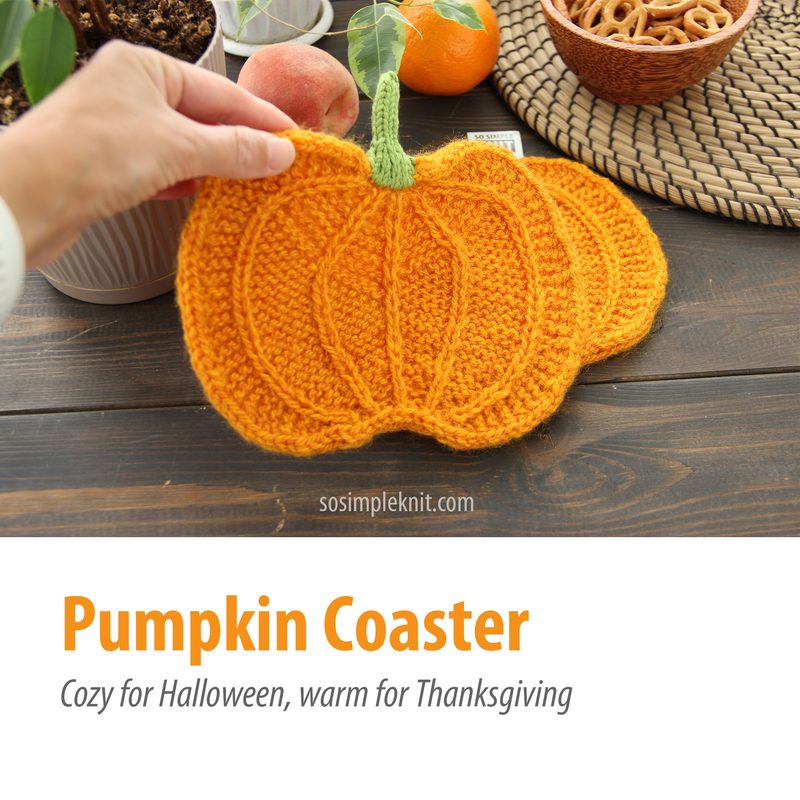A hand holds an orange knitted pumpkin coaster, featuring a textured, ribbed design with green stems. The coaster is set against a wooden table with a woven placemat underneath, and various autumn-themed items in the background, including an orange, an apple, and a bowl of pretzels. The image is framed to focus on the coaster, with a warm and inviting ambiance, highlighting the seasonal theme.