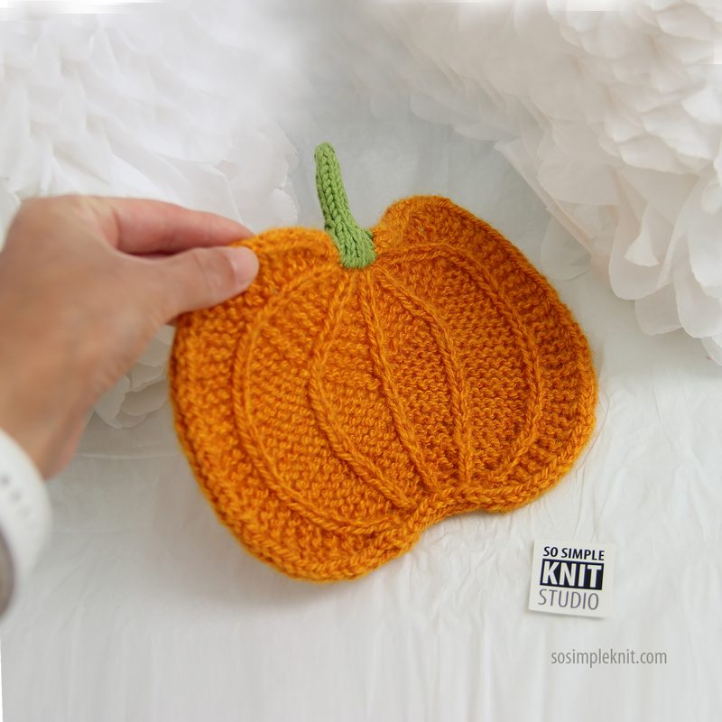 hand holding a knitted pumpkin in vibrant orange yarn, featuring textured knit lines and a green knitted stem on top. The pumpkin is placed against a soft white background, with a subtle shadow cast underneath. The lower right corner shows a small label with "So Simple Knit Studio" printed on it.