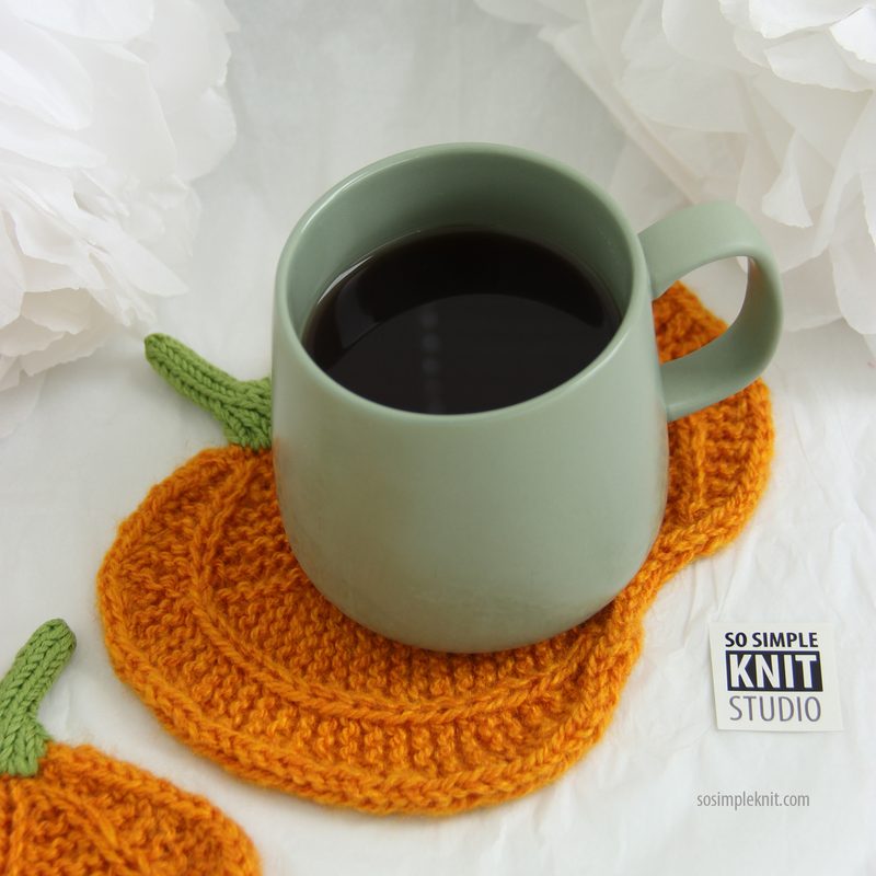 A green ceramic cup filled with dark liquid sits on an orange knitted coaster shaped like a pumpkin, which features green knit leaf details, against a light background with soft white paper flowers in the backdrop. The composition highlights the contrasting colors and textures of the cup, coaster, and flowers.