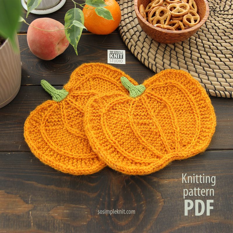 The image features two knitted pumpkin-shaped coasters in vibrant orange, with green stem details. They are placed on a dark wooden surface alongside a woven basket containing pretzels and an orange. Soft light highlights the texture of the knitted pumpkins, emphasizing their detailed stitches and the cozy atmosphere of the setting. The text "Knitting pattern PDF" is displayed in the bottom right corner.