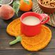The image shows a vibrant orange pumpkin-shaped knitted coaster with a green knitted stem, placed on a wooden surface. A bright red mug filled with a light-colored drink sits on the coaster. Surrounding the mug are a peach, an orange, and a bowl of pretzels, creating a cozy, seasonal setting. A few green leaves are partially visible in the background, adding to the natural feel of the composition.