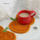 The image features a knitted pumpkin coaster in vibrant orange, shaped like a pumpkin with a green stem, resting on a soft white background. A small red ceramic cup filled with a light-colored beverage sits on top of the coaster, creating a cozy scene. Fluffy white tissue paper decoratively frames the composition, enhancing the softness of the overall aesthetic. A logo for "So Simple Knit Studio" is visible near the bottom right corner.
