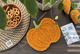 A knitted pumpkin coaster in vibrant orange is placed on a dark wooden table alongside a woven placemat and a small bowl of pretzels. Nearby, a printed knitting pattern brochure featuring floral illustrations is visible, and a small tag with "So Simple Knit Studio" is displayed. Green leaves from a plant hang slightly over the scene, adding a natural touch.