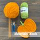 The image displays a vibrant orange ball of yarn next to a green skein, with a completed knitted pumpkin motif placed next to them. Above, a pair of knitting needles and a small pair of scissors are positioned alongside a stitch marker and a blue crochet hook. The background consists of a dark wooden surface, with the text "Materials & tools" prominently displayed at the bottom in white and gray.