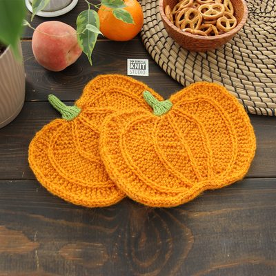 Two knitted orange pumpkins with green stems. Knitting pattern for a pumpkin-shaped mug coaster