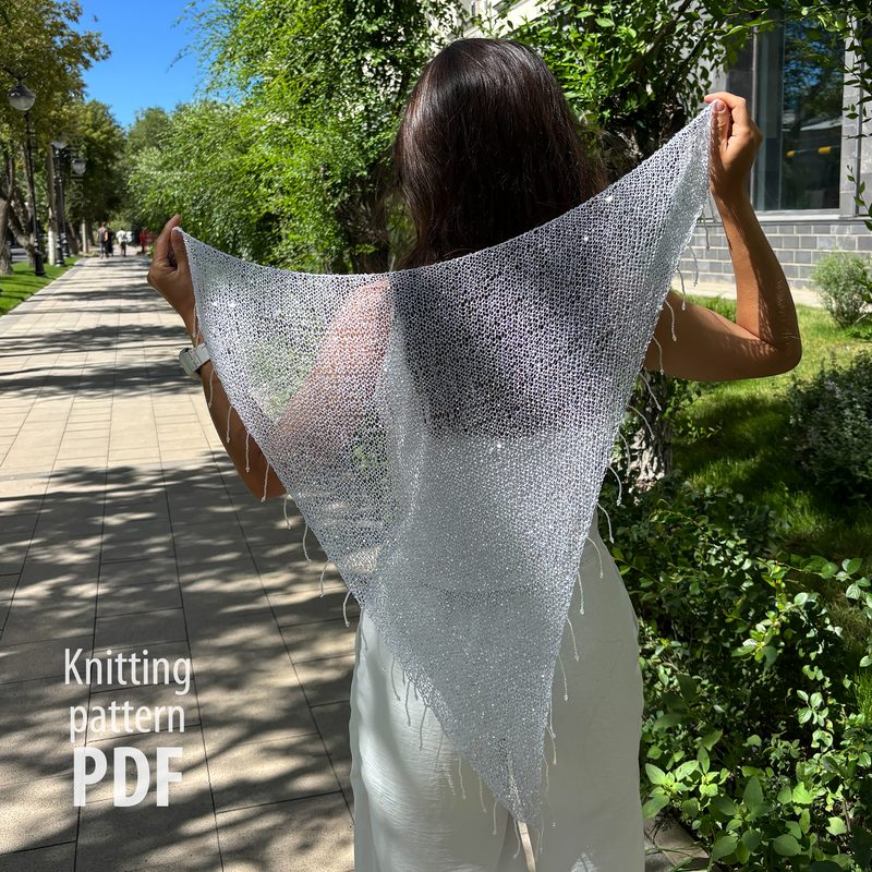 A woman stands outdoors, facing away, holding up a shimmery, silver sequin shawl that is triangular in shape. The shawl features a sheer, sparkling design with fringes along the edges. She is dressed in a light-colored outfit, and the background shows a tree-lined sidewalk with greenery and a building partially visible on the left. The text "Knitting pattern PDF" is displayed prominently on the left side of the image.