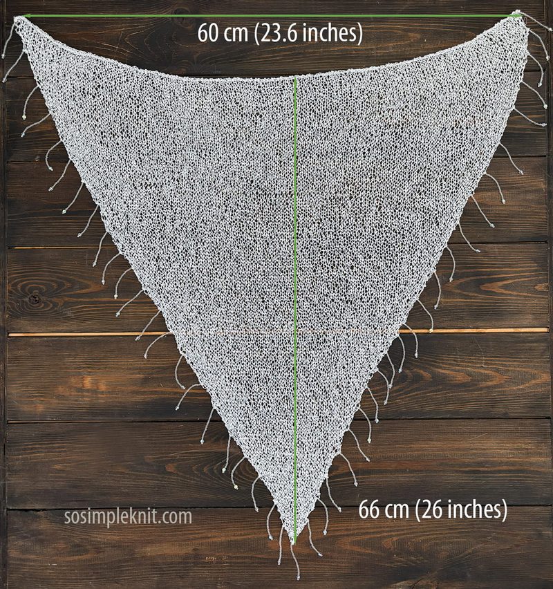 A triangular sequin shawl is displayed against a dark wooden background. The shawl is primarily white, embellished with shiny silver sequins that reflect light. The edges are finished with loose, delicate threads that hang down slightly. Measurement lines indicate the dimensions of the shawl, with the width marking 60 cm (23.6 inches) and the height marking 66 cm (26 inches). The overall composition is focused on showcasing the shawl's design and texture.