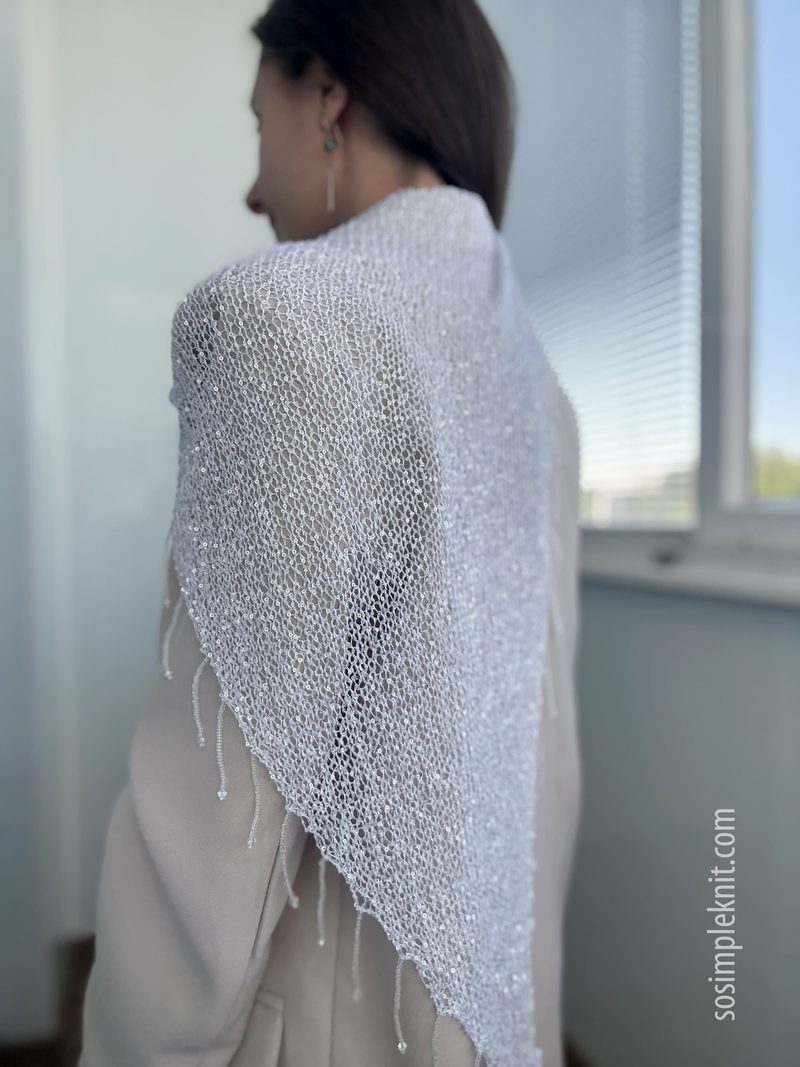 A woman is modeling a shimmering silver sequin shawl draped over her shoulders, showcasing its delicate knit texture. The shawl features a triangular shape with decorative fringes at the bottom. The background is a softly lit room with large windows, creating a bright atmosphere. The woman is wearing a light-colored top and is partially turned away from the camera, emphasizing the elegant draping of the shawl.