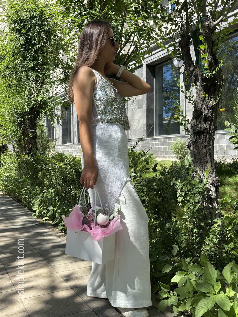 A woman stands outdoors along a tree-lined walkway, wearing a sleeveless sequined top that reflects sunlight, paired with flowy white trousers. She holds a small white handbag with pink accents in one hand and poses with a confident posture, her hair falling over one shoulder. The background features greenery and a modern building, adding a vibrant and stylish context to the image.
