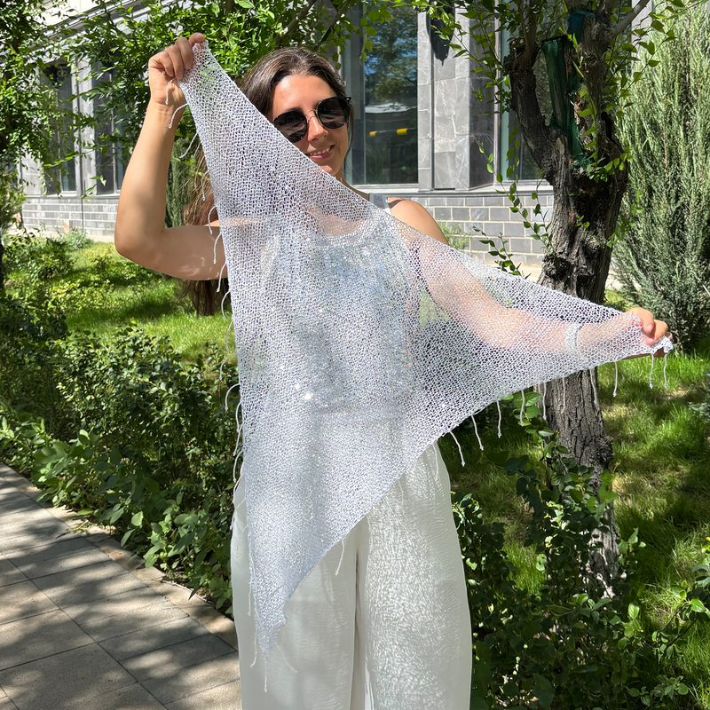 A woman stands outdoors holding a large, triangular sequin shawl that sparkles in the sunlight. The shawl is made of sheer, silvery material with intricate sequin details. She is dressed in light-colored, flowy pants and a tank top, wearing sunglasses. In the background, there are green trees and a building with large windows. To the left of the image, there is text that reads "Knitting pattern PDF." The overall scene is bright and inviting, with a natural setting.