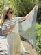 A woman stands outdoors holding a triangular sequin shawl, showcasing its shimmering texture. She wears a striped short-sleeve shirt and light-colored trousers, with long hair flowing down her back. The shawl is predominantly silver with a loose-knit design, allowing light to reflect off the sequins. The setting features greenery in the background, indicating a sunny day and a casual environment.