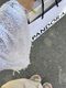 The image shows a close-up of a person's arm wearing a white, knitted shawl with sequins that shimmer in the light. The edge of the shawl has a loose, fringed texture. The hand is holding a black and white shopping bag marked with the word "Pandora," and a part of a book, visible above the bag, has a yellow bookmark protruding from it. The background is blurred, showing a textured surface, possibly pavement, suggesting the setting is outdoors. The person's feet, clad in white sandals, are also p
