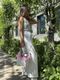 A woman stands outdoors along a tree-lined walkway, wearing a sleeveless sequined top that reflects sunlight, paired with flowy white trousers. She holds a small white handbag with pink accents in one hand and poses with a confident posture, her hair falling over one shoulder. The background features greenery and a modern building, adding a vibrant and stylish context to the image.