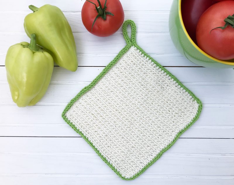 The image features a single square knitted dishcloth with a white body and a green border. It is placed on a light, textured surface and is accompanied by two green bell peppers and two red tomatoes, arranged beside a round green bowl holding the tomatoes. The dishcloth is positioned at a slight angle, showcasing its texture.