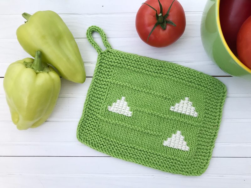 A bright green knitted dishcloth featuring three white mountain shapes is positioned on a white wooden surface. To the left, there are two yellow bell peppers, and to the right, a vibrant green bowl containing red tomatoes. The arrangement highlights the dishcloth and surrounding produce.