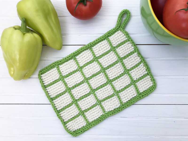 A knitted dishcloth with a checkered pattern of green and white is positioned on a white wooden surface, accompanied by two green bell peppers and a bowl of red tomatoes. The dishcloth features a loop for hanging and has a bright, textured appearance, with the green edges framing the checkered design. The overall composition is colorful and well-balanced, highlighting the dishcloth and fresh produce.