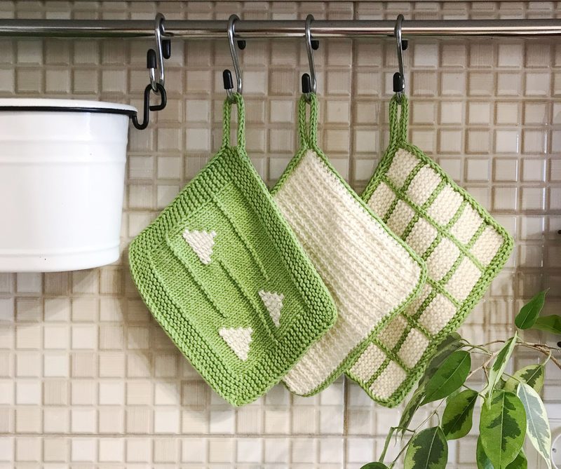 Three knitted dishcloths hang from a metal rod in a kitchen setting. The left dishcloth is green with a textured pattern and white triangles. The middle dishcloth is solid cream-colored with a smooth surface. The right dishcloth features a grid design in shades of green and cream. Above the dishcloths, there is a white bucket hanging, and the background consists of light beige tiles with a subtle grid pattern. A green plant is partially visible in the foreground.