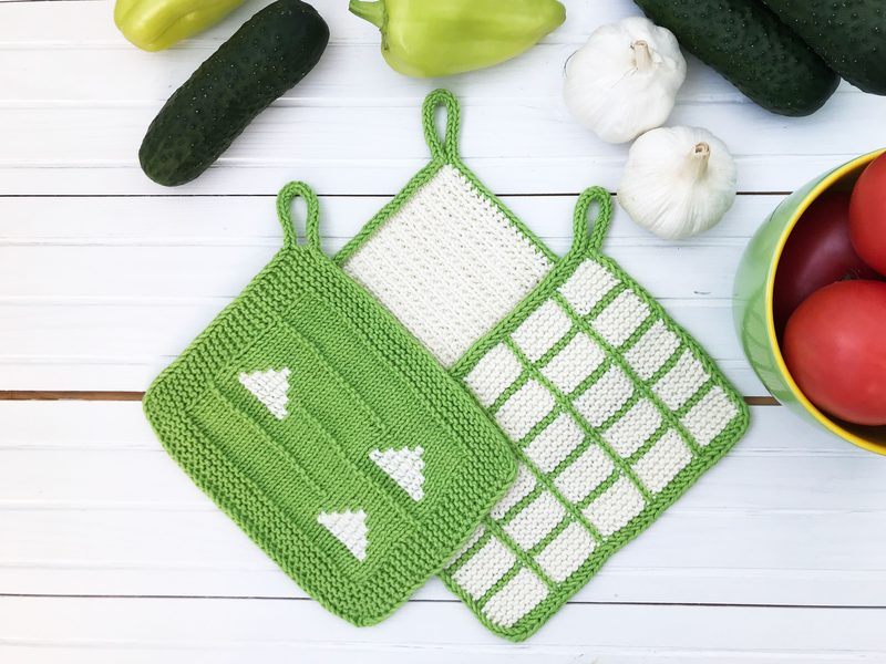The image displays a set of three knitted dishcloths arranged on a white wooden surface, with two dishcloths in bright green featuring white patterns of trees and a grid, and one in cream with a green border. Surrounding the dishcloths are fresh cucumbers, a yellow bell pepper, garlic cloves, and a bowl of red tomatoes, adding a vibrant contrast to the scene. The overall composition highlights the dishcloths' texture and colors against a rustic background.