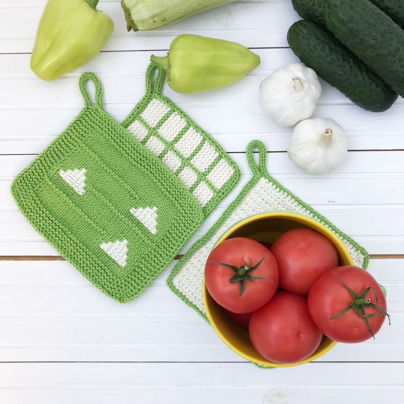 Image of three knitted dishcloths arranged on a white wooden surface. Two dishcloths are green with white patterns: one features a grid design, and the other has white triangles. The third dishcloth is white with a green border. Surrounding the dishcloths are fresh vegetables including green bell peppers, cucumbers, garlic, and a bowl of red tomatoes in the foreground. The colors are vibrant and emphasize a fresh, homey kitchen setting.