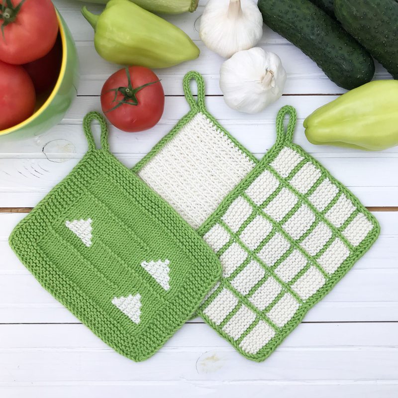 A set of three knitted dishcloths displayed on a white wooden surface. The dishcloths include a green one with white triangle designs, a plain white one, and a green and white checkered one. Surrounding them are fresh tomatoes, green peppers, and cucumbers, with a yellow bowl holding more tomatoes in the top left corner. The overall composition features a bright and vibrant color palette, emphasizing the knitted texture of the dishcloths against the natural produce.