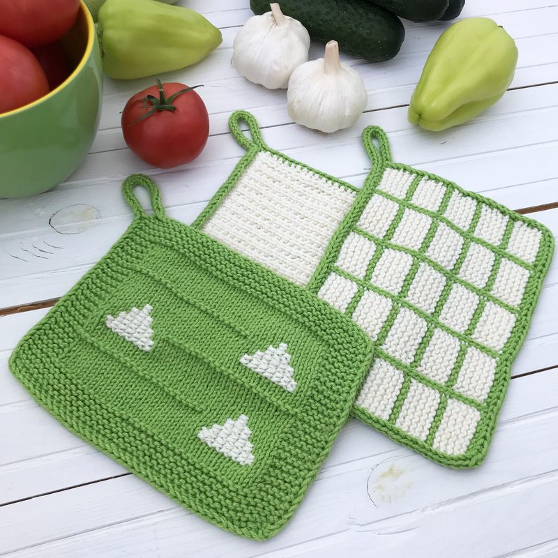 A set of three knitted dishcloths is displayed on a white wooden surface. The dishcloths feature a vibrant green border with a design of white hearts on one, a textured knit pattern on another, and a checkered grid on the third. Surrounding the dishcloths are fresh vegetables, including red tomatoes, green cucumbers, and garlic, which add a pop of color to the scene. The overall composition is bright and inviting, showcasing the dishcloths prominently against the light background.