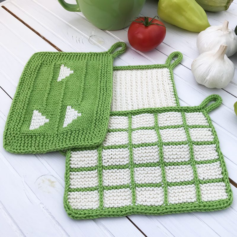 The image features a set of three knitted dishcloths, showcasing a green and white color scheme. One dishcloth has a row of green triangles on a white background, while another displays a grid pattern with alternating green and white squares. The third cloth is solid green with a border and a hanging loop. The dishcloths are placed on a light wooden surface, accompanied by a green mug and colorful vegetables, enhancing the homey setting.