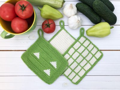 The image features a set of three knitted dishcloths arranged on a white wooden surface, surrounded by fresh vegetables. The dishcloths are in shades of green, one with white arrows, another with a textured pattern, and the third with a grid design. In the background, there is a bowl of red tomatoes and various green vegetables, including cucumbers, garlic, and bell peppers, adding color and context to the kitchen setting.
