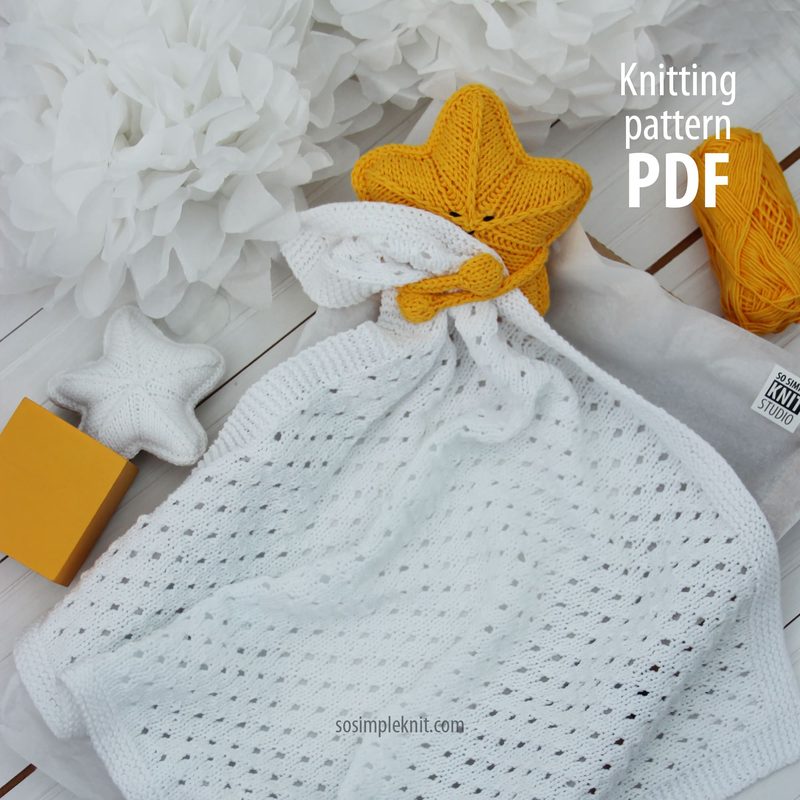 A white knitted security blanket with an open stitch pattern is laid flat on a light wooden surface. At one corner, there is a large yellow star-shaped head. Nearby, a smaller white star and a yellow yarn ball are visible. White paper flowers create a soft background, and a small orange box adds a pop of color. The text "Knitting pattern PDF" is displayed in the upper right corner.