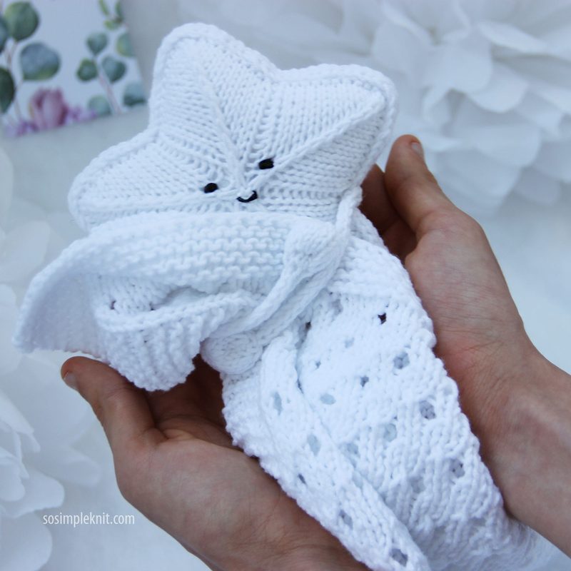 A pair of hands gently holds a white knitted security blanket shaped like a star, featuring a smiling face with two black button-like eyes. The star's body has a textured knit pattern, while the blanket has a lacy design along the edges. The background is softly blurred, with light colors and floral motifs, creating a serene, cozy atmosphere.
